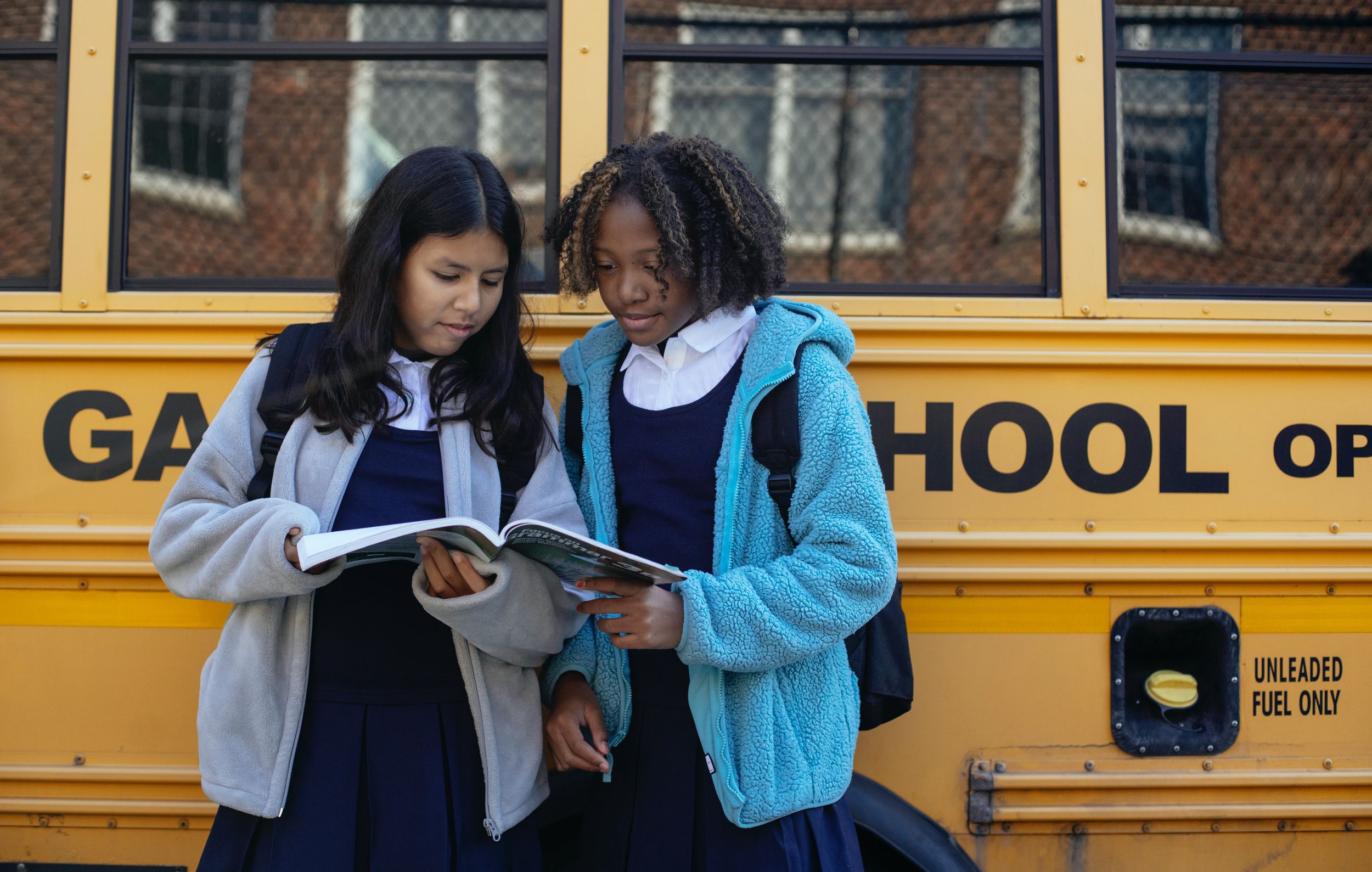 Two girls in uniforms stand outside of a yellow school bus.
