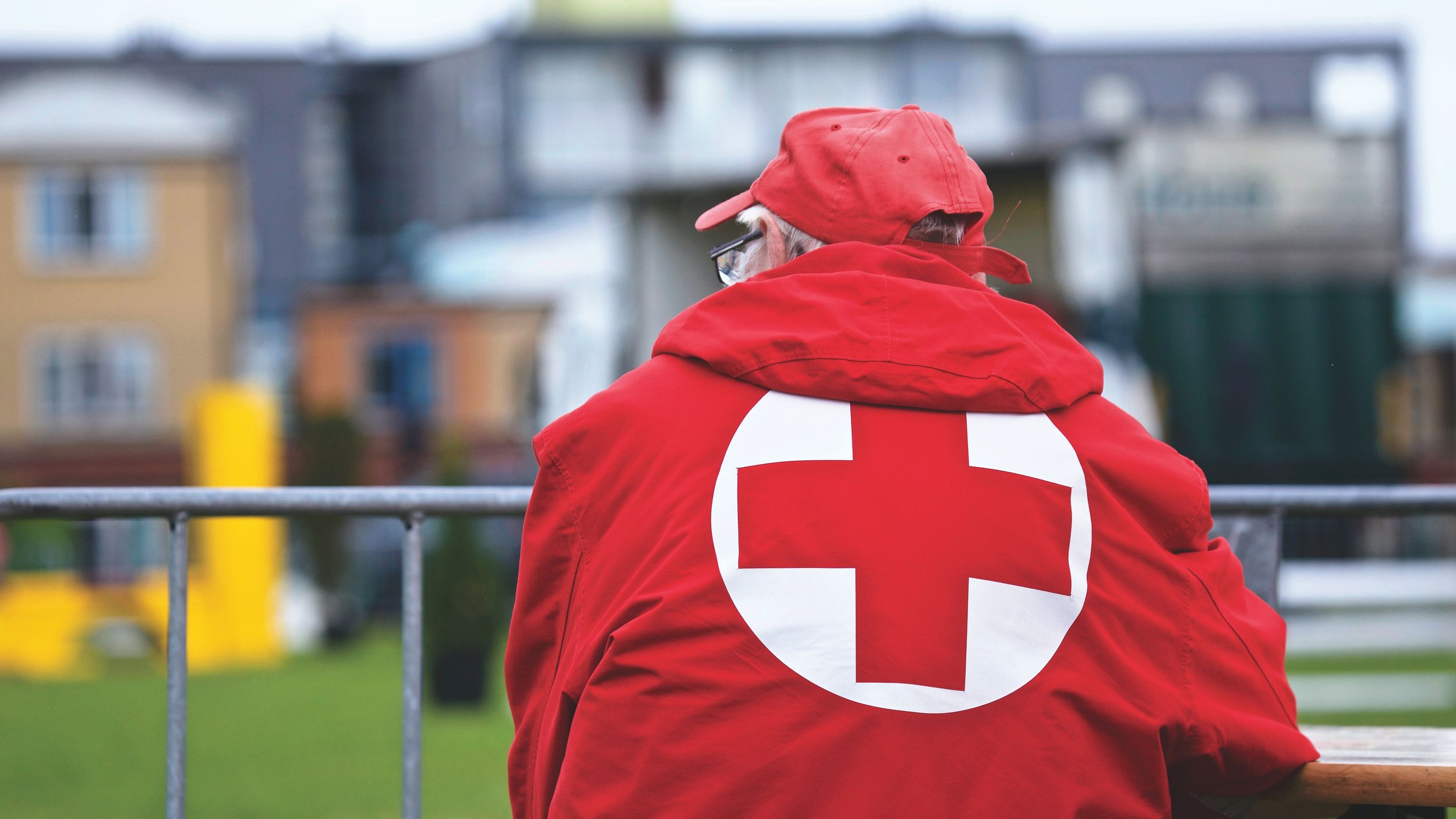 Man in a red cross jacket stands by a rail.